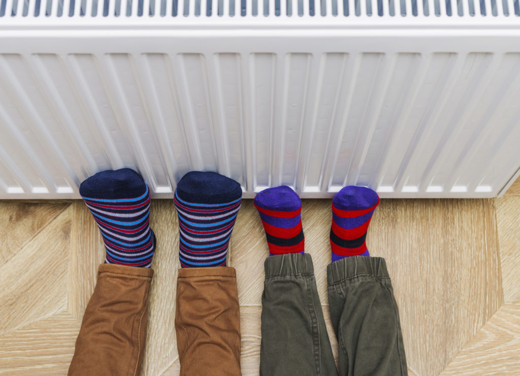 Woman and child wearing colorful pair of socks warming cold feet in front of heating radiator. Gas heater at home. Selective focus. Warming feet on a radiator